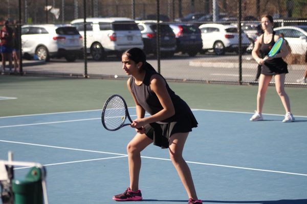 Freshman Rhea Sethi stands in a high squat as one of her opponents serves the ball from the opposite side of the court.
