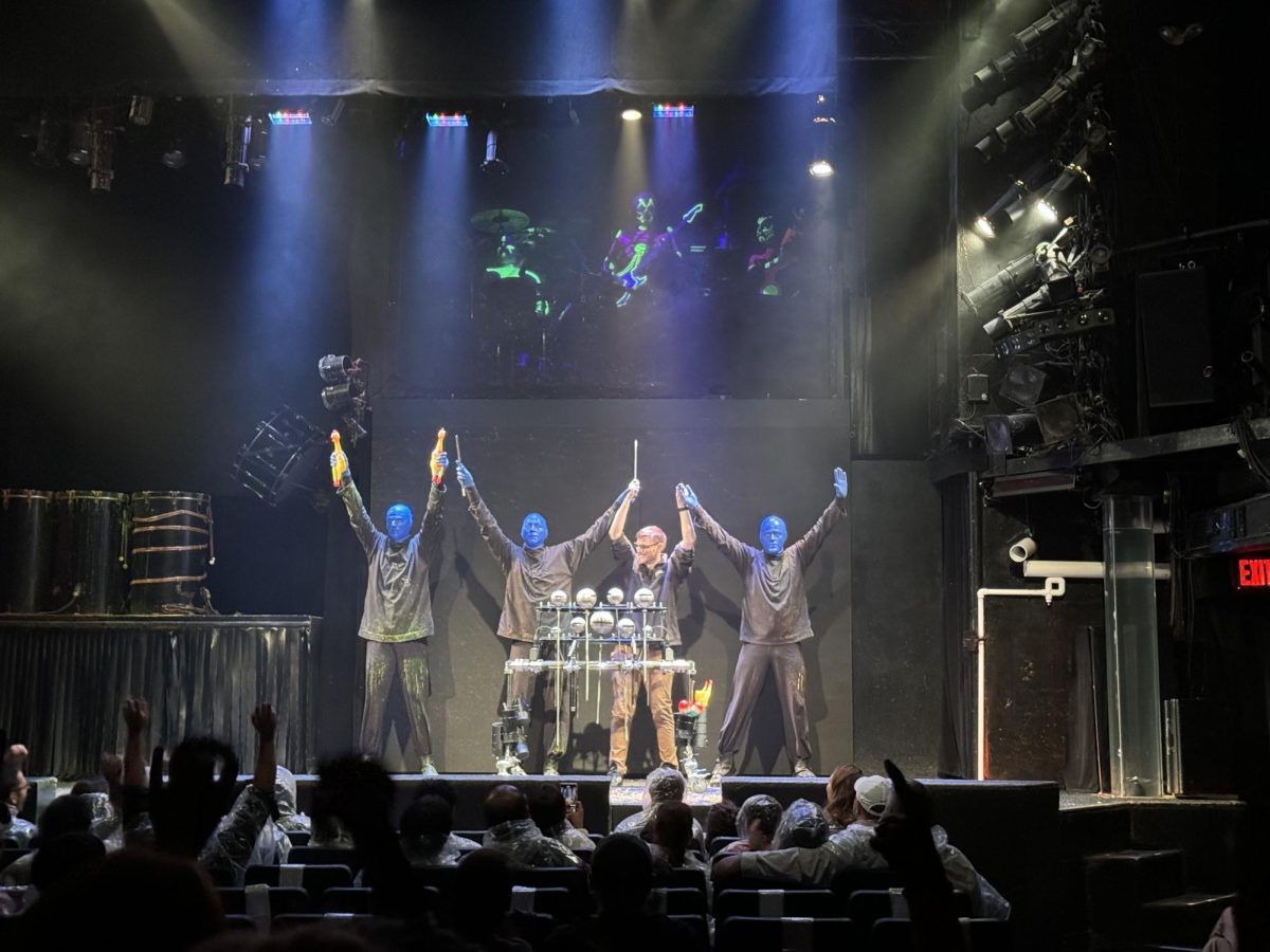 After bringing an audience member up to the stage and performing with them, the Blue Men and audience member raise their arms up in celebration. 