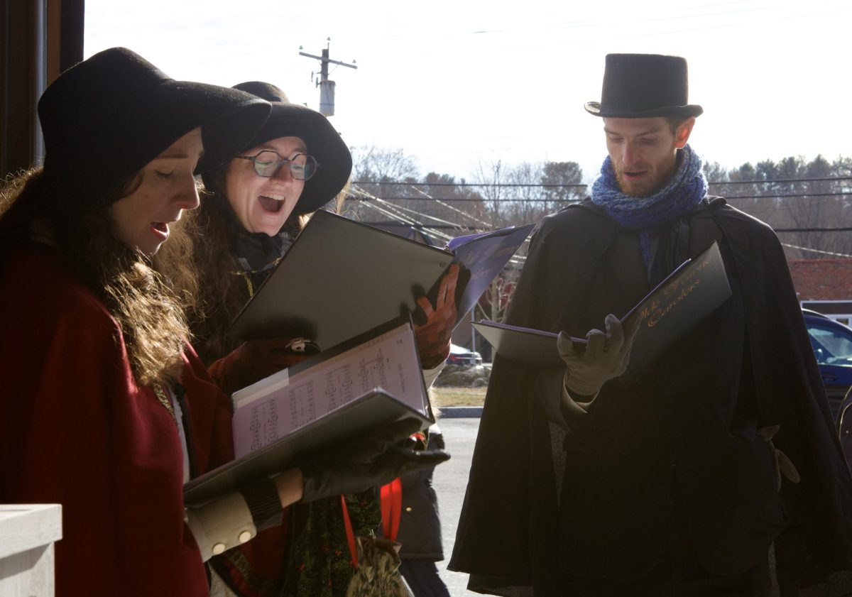 A group of three carolers sing holiday tunes. The group of carolers walked around the market and sang to the shoppers at different locations. They sang many festive songs including "Rudolph, the Red-Nosed Reindeer" by Gene Autry and The Pinafores.