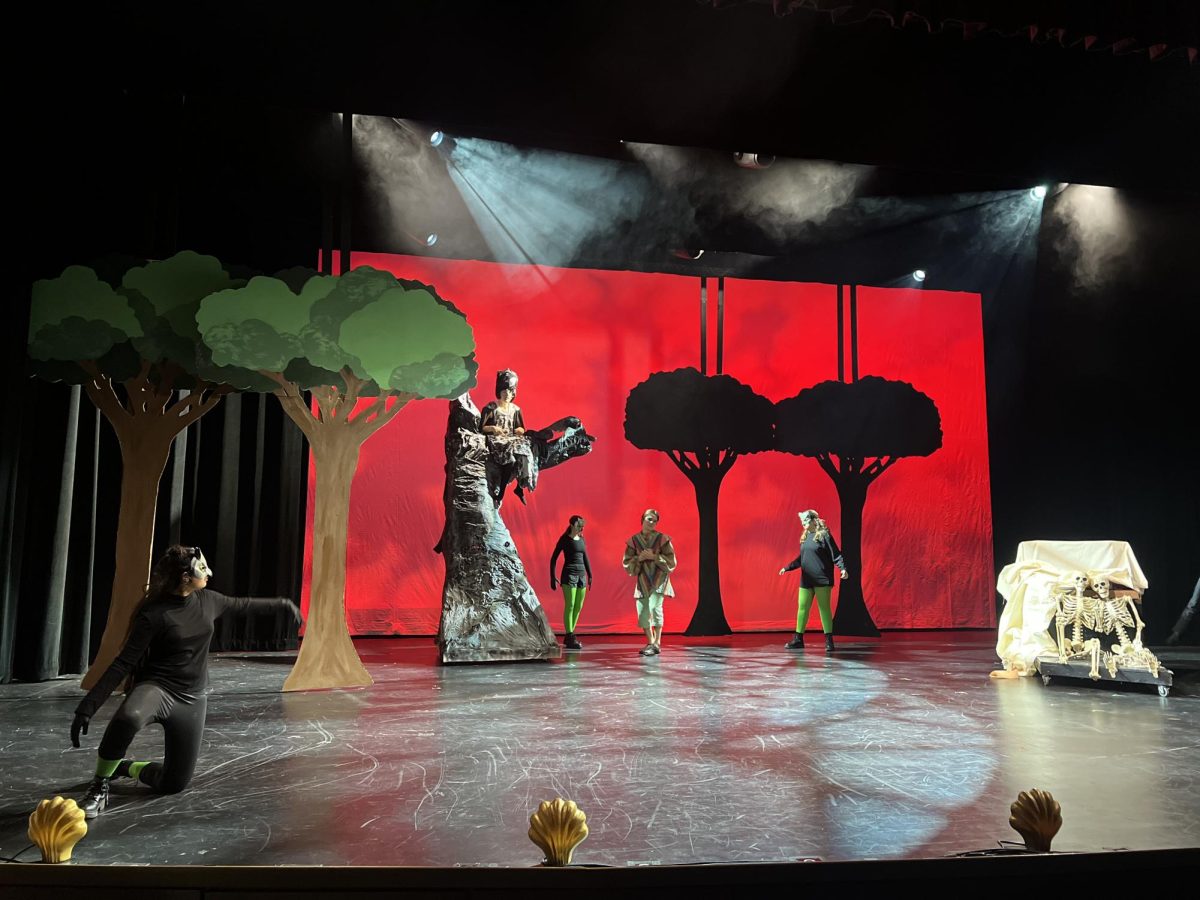 Senior Jules Eggers plays an oracle from Pythio while sitting in a papier-mâché tree. The cast acts out a suspenseful scene against an eerie backdrop.