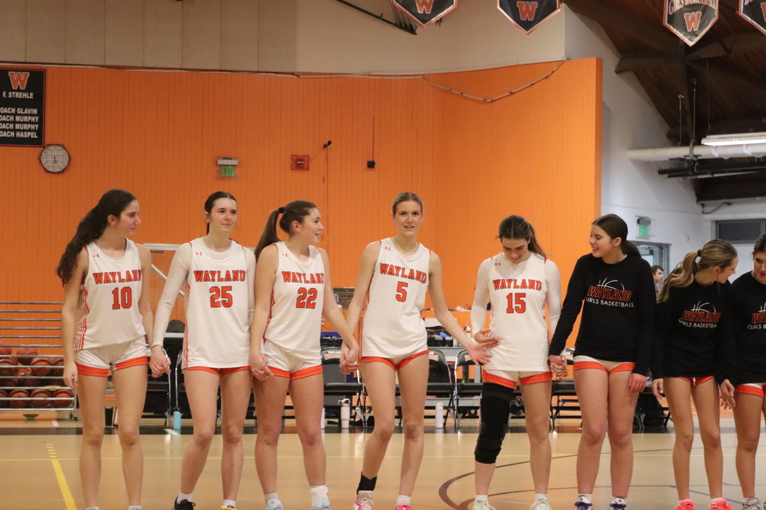 The girls varsity basketball team joins hands for the national anthem. The anthem is sung before each varsity game, and teams join hands and face the flag in respect.